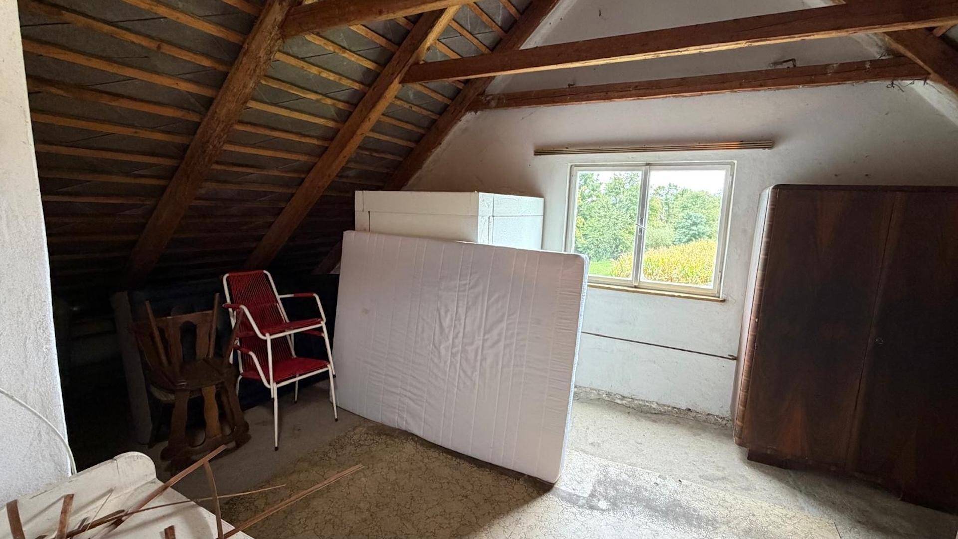 Dachbodenraum mit sichtbaren Holzbalken, Fenster und Stauraum, unfertiger Zustand.