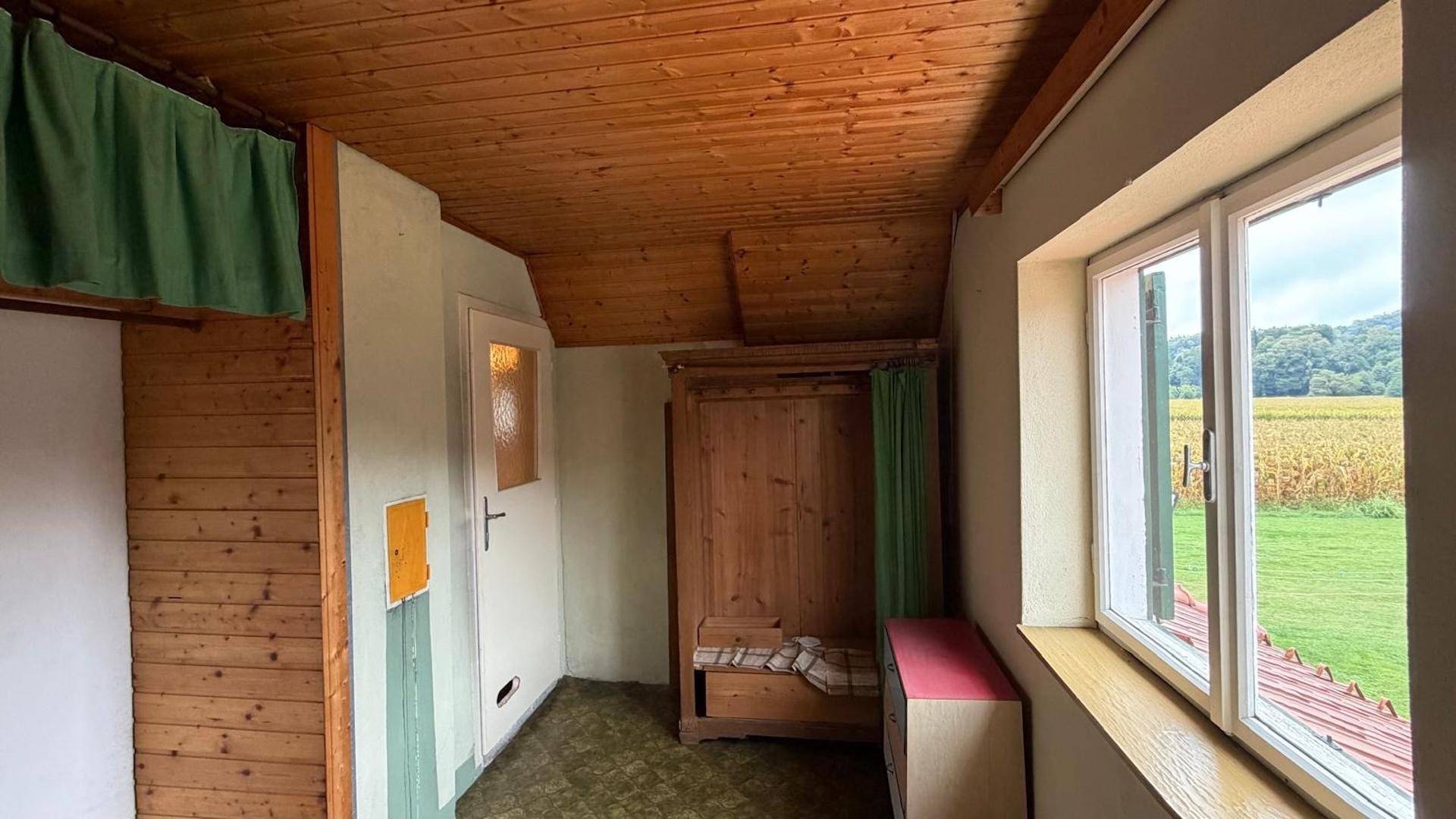 Zimmer mit Holzvertäfelung, Fenster mit Blick ins Grüne und einfacher Einrichtung.