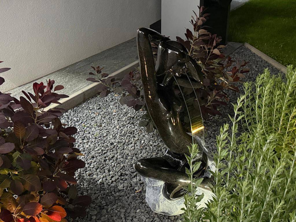 Detailaufnahme einer modernen Gartenskulptur umgeben von Zierkies und Pflanzen im Abendlicht.