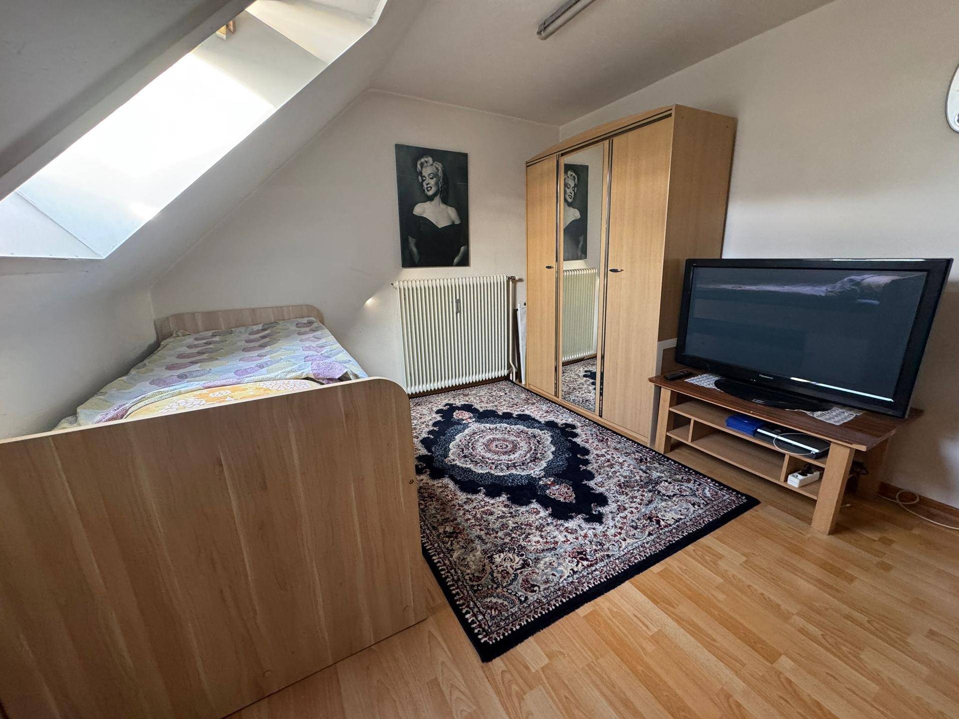 Helles Dachgeschoss-Schlafzimmer mit Einzelbett, Kleiderschrank und Blick aus dem Dachfenster.