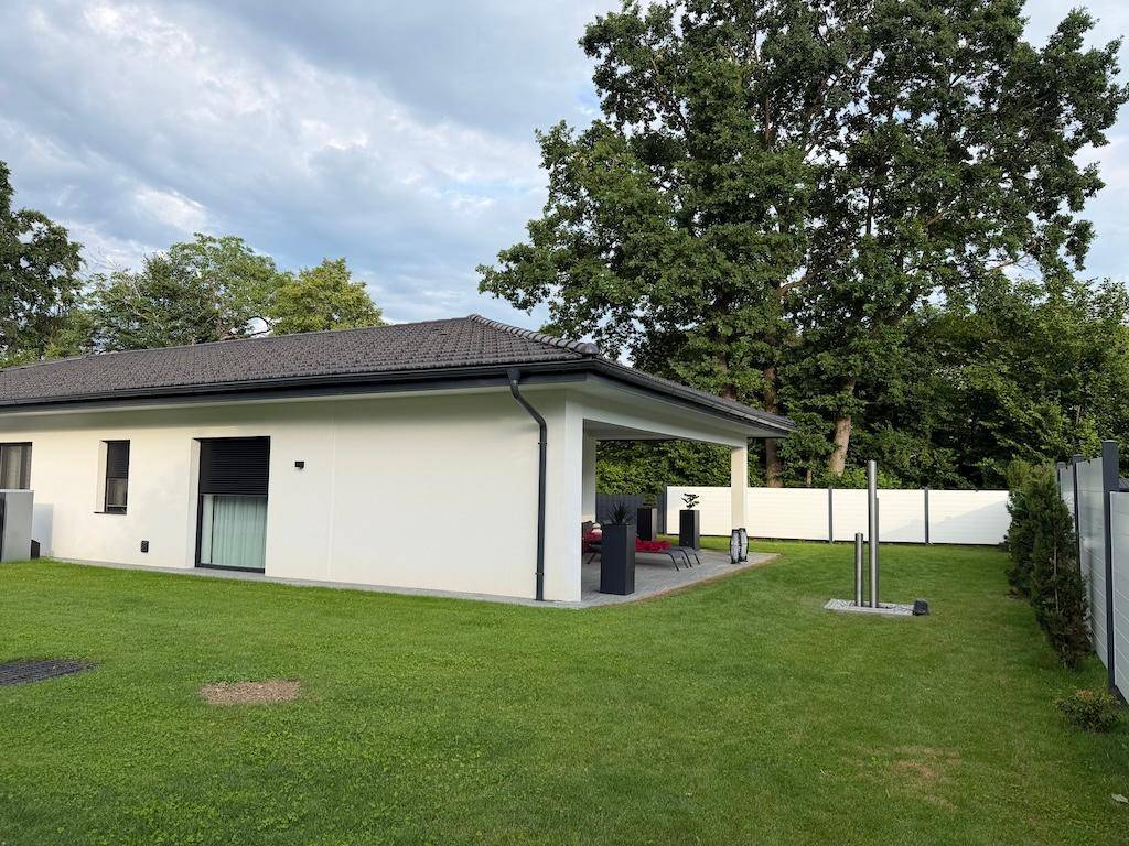 Seitenansicht des Hauses mit einer überdachten Terrasse, großem Rasen und einem modernen Zaun.