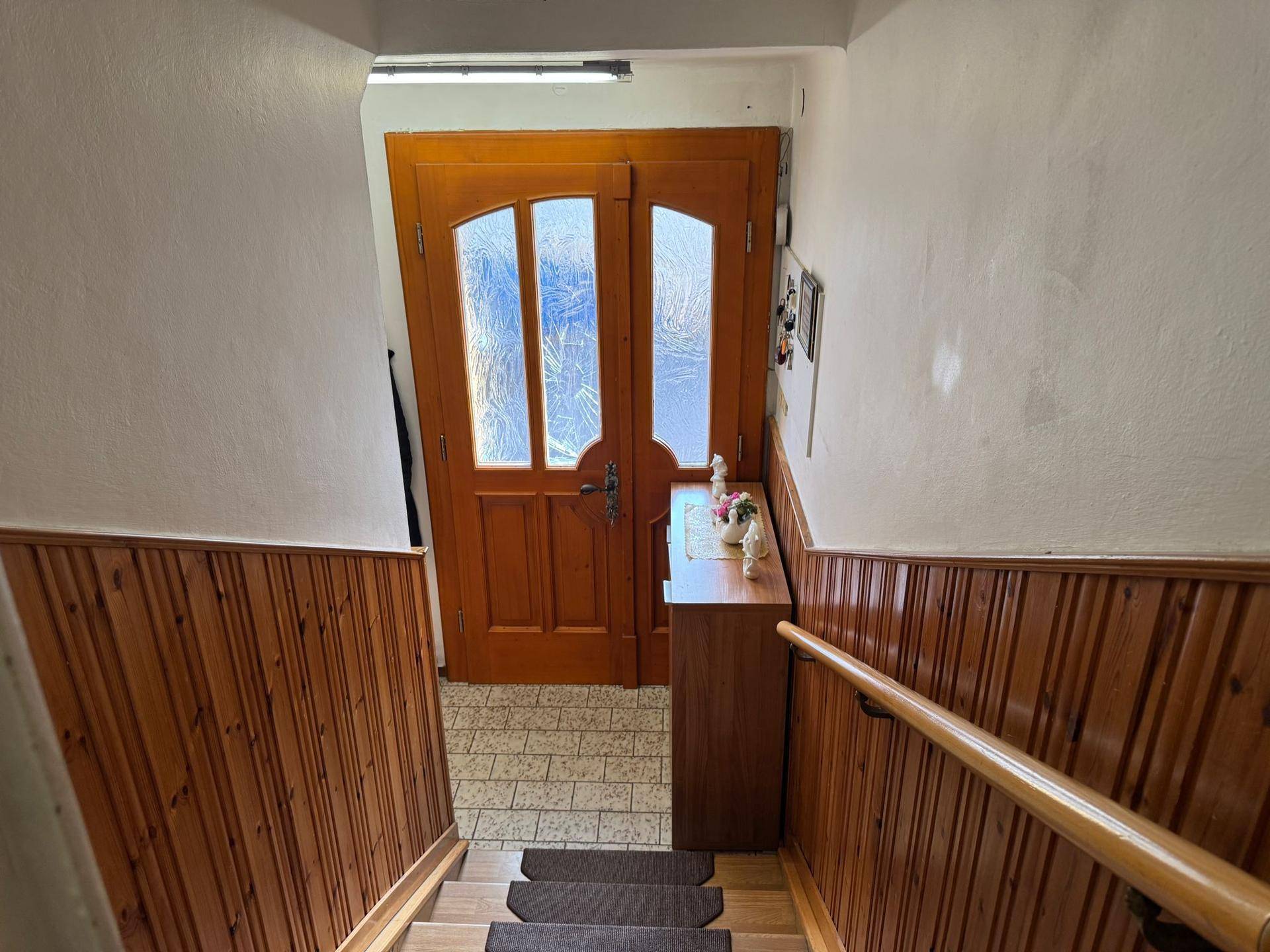 Treppenhaus mit Holzvertäfelung und gefliestem Eingangsbereich, der zu einer Holztür führt.
