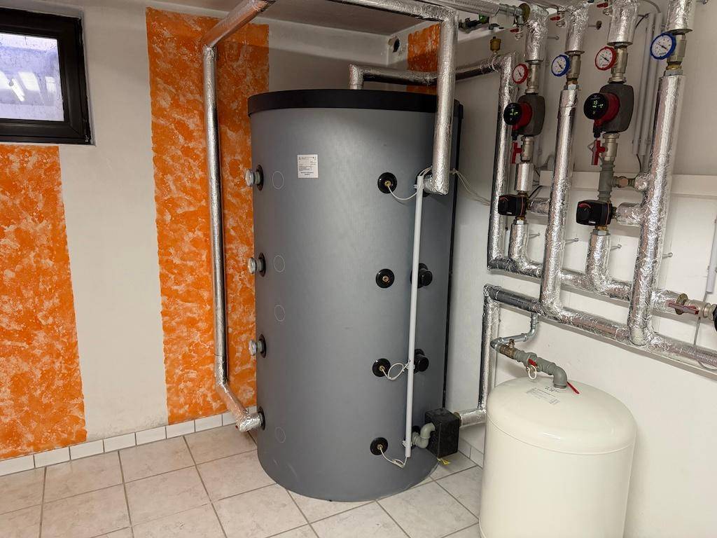 Großer Warmwasserspeicher und Heizungsrohre in einem technischen Raum mit Fliesenboden.