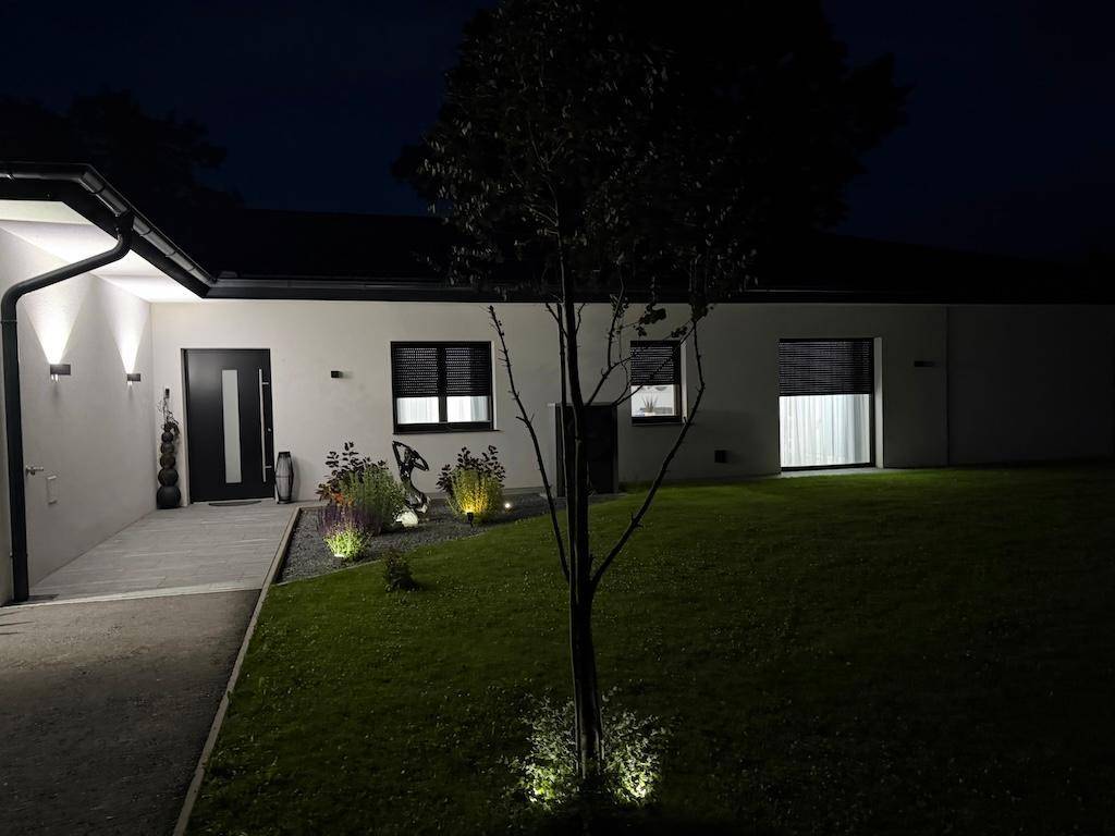 Eingangsbereich des Hauses bei Nacht mit dezenter Beleuchtung und kleinen Pflanzenbeeten.