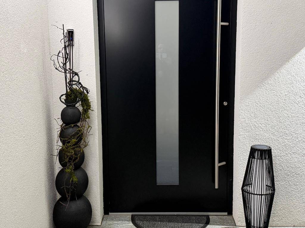 Moderner Hauseingang mit schwarzer Tür, seitlichem Glaseinsatz und stilvoller Dekoration.