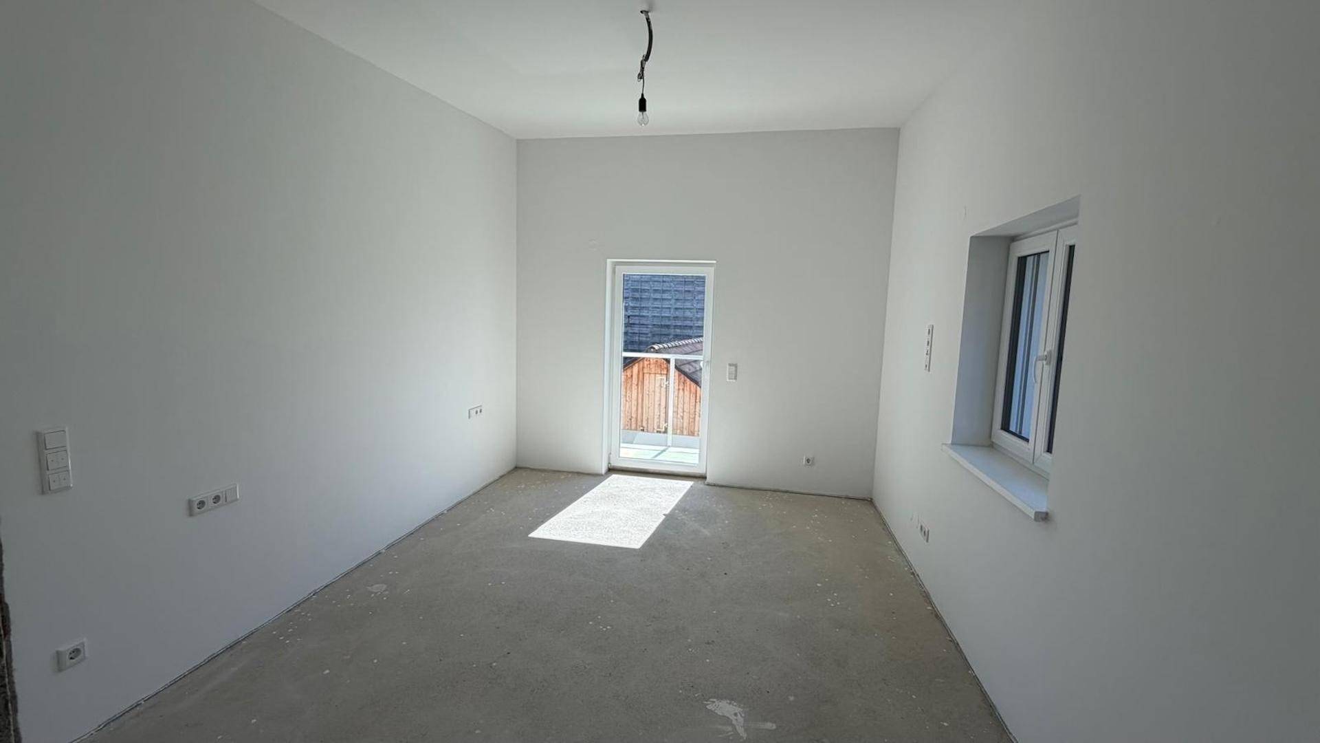 Leerer Raum im Rohbau mit Tür zum Außenbereich und einem Fenster, das viel Tageslicht hereinlässt.