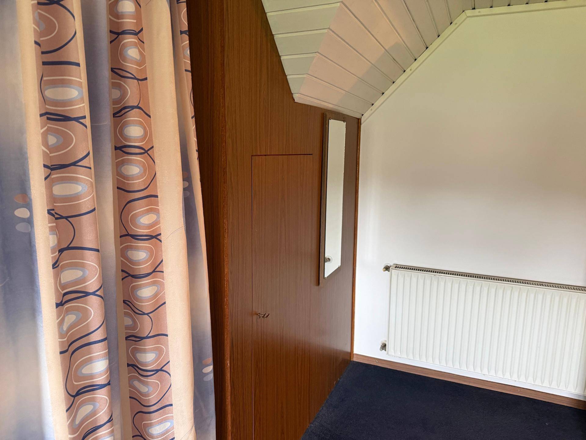 Schlafzimmer mit Dachschräge, Einbauschrank und Heizkörper, teilweise verdeckt von Vorhängen.