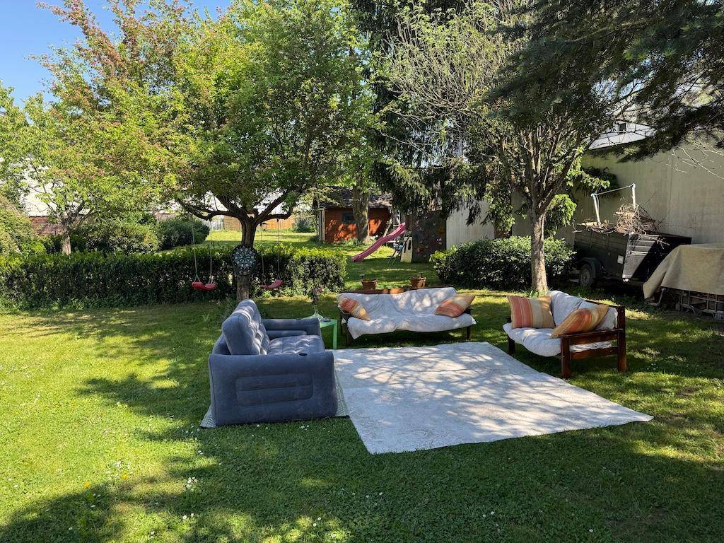 Großer grüner Garten mit Sitzgelegenheiten, Rasenfläche und Bäumen im Sonnenlicht.