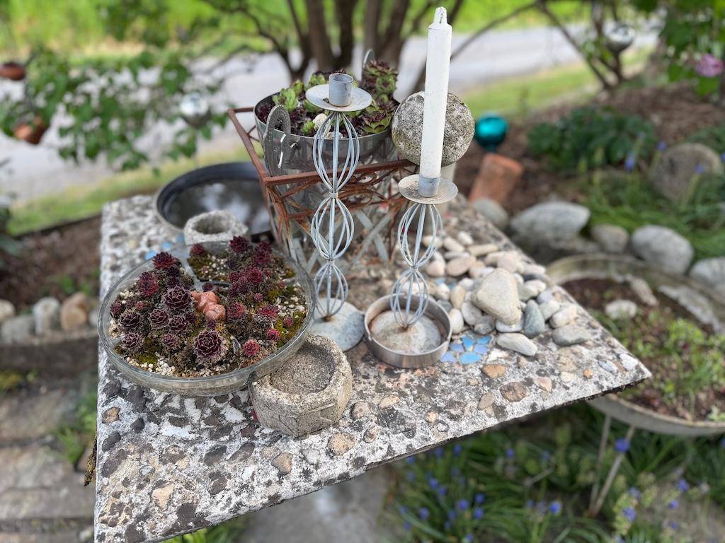 Dekorative Gartengestaltung mit Sukkulenten, Kerzenständern und Steinen auf einem Betontisch.