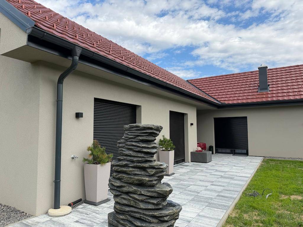 Gepflasterte Terrasse mit modernem Steinfbrunnen und schwarzen Rollläden an den Fenstern des Hauses.
