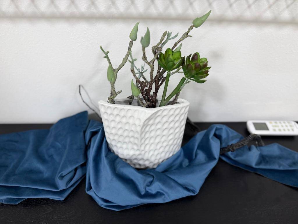Nahaufnahme einer weißen, strukturierten Vase mit grünen Pflanzen und blauer Stoffdekoration.