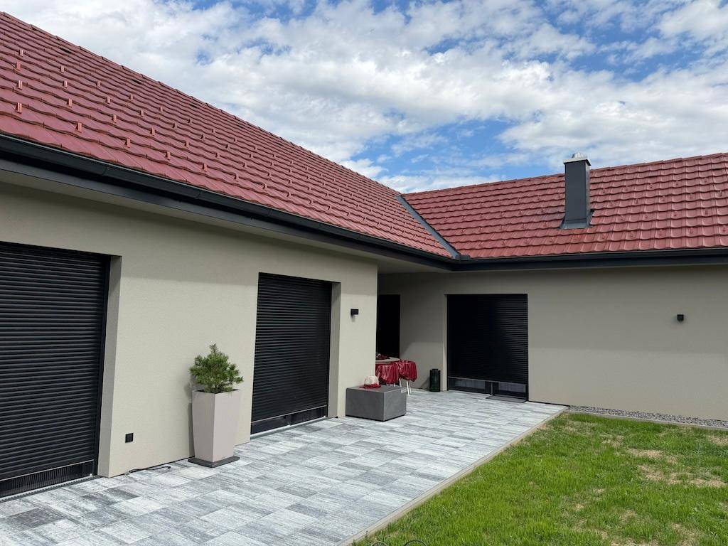 Gepflasterte Terrasse mit kleinem Esstisch und schwarzen Rollläden an den Fenstern des modernen Hauses.