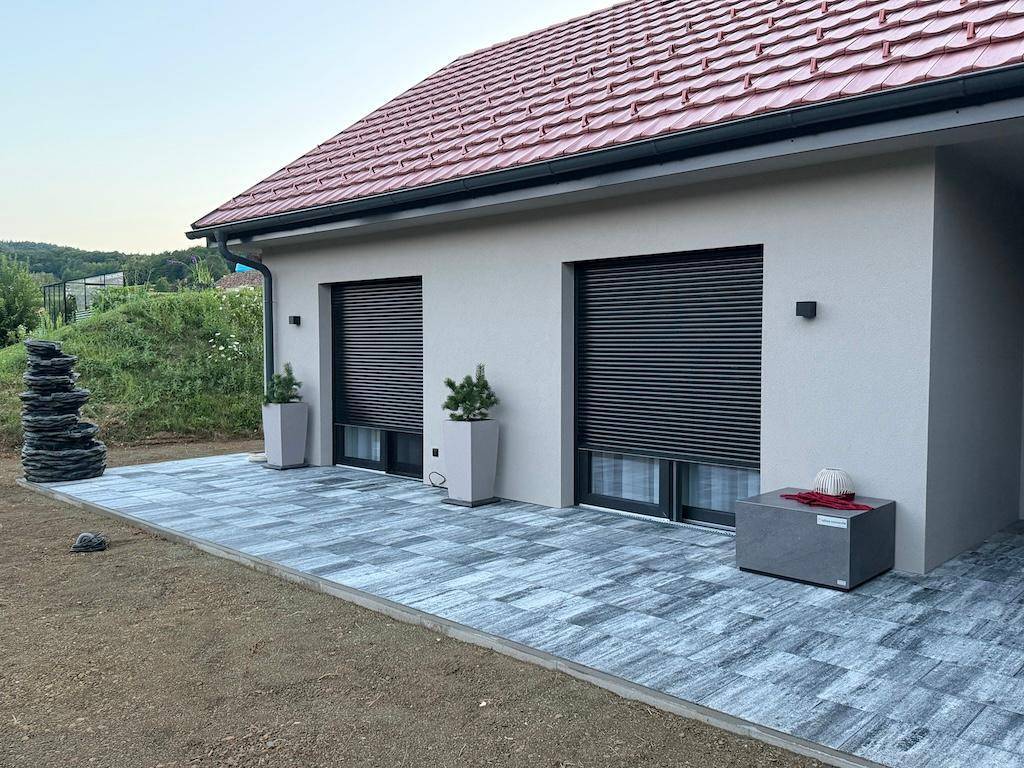 Geräumige Terrasse mit Fliesenboden und geschlossenen Rollläden, umgeben von junger Bepflanzung.