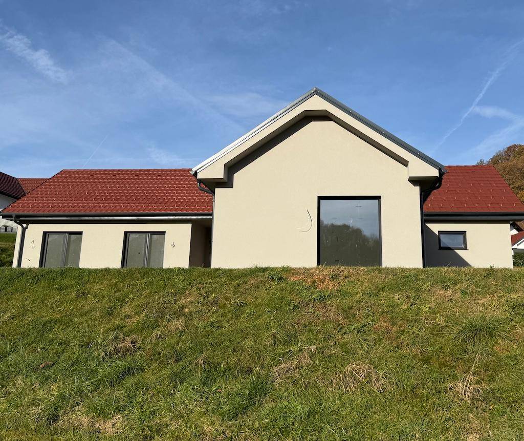 Modernes Einfamilienhaus mit heller Putzfassade und rotem Dach, umgeben von einer grünen Wiese.