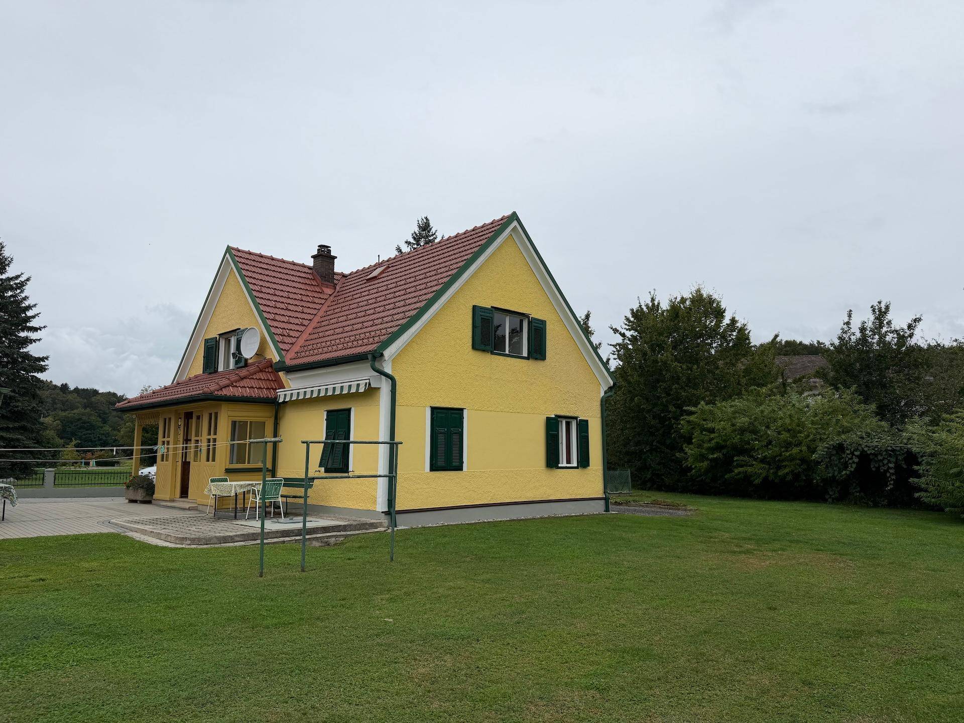 Ansicht des Hauses mit gelber Fassade, rotem Dach und einem grünen Gartenbereich mit Sitzgelegenheiten.