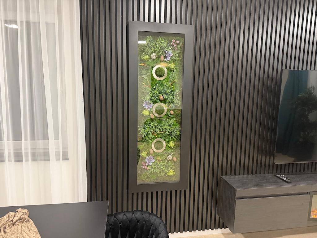 Moderne Wanddekoration mit vertikalen Holzlamellen und einem beleuchteten Pflanzenbild im Wohnbereich.