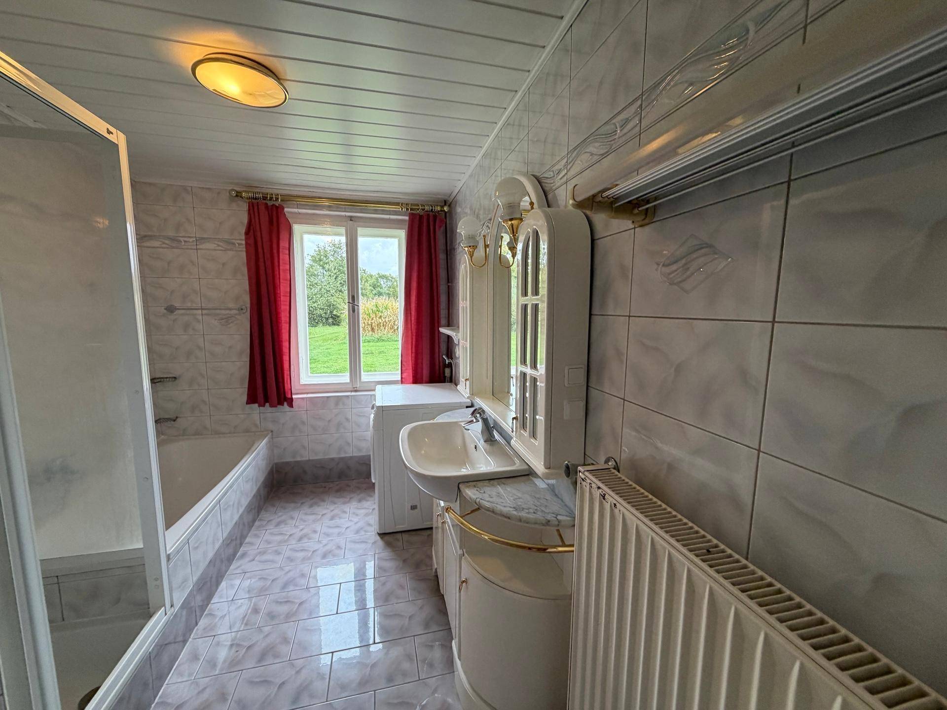 Helles Badezimmer mit Badewanne, Waschbecken, Waschmaschine und Fenster mit Blick ins Grüne.