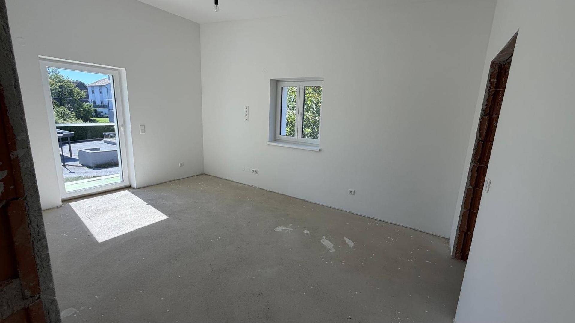Geräumiger, unfertiger Raum mit Terrassenzugang und Fenster, bereit für den Ausbau.