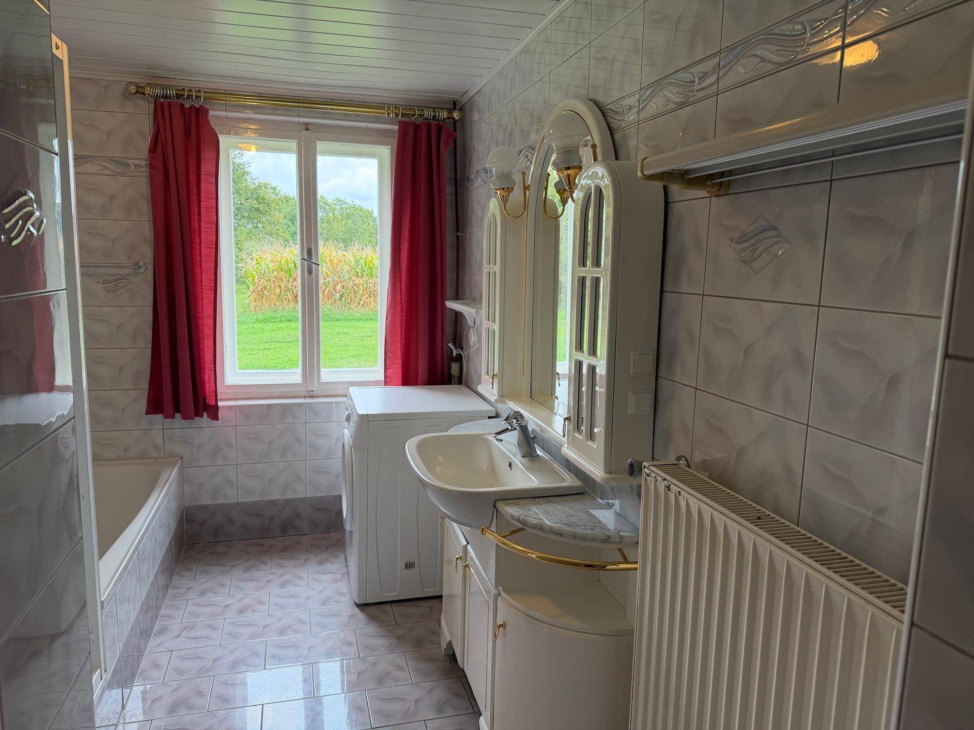 Helles Badezimmer mit Badewanne, Waschbecken, Spiegelschrank und Fenster mit Blick ins Grüne.