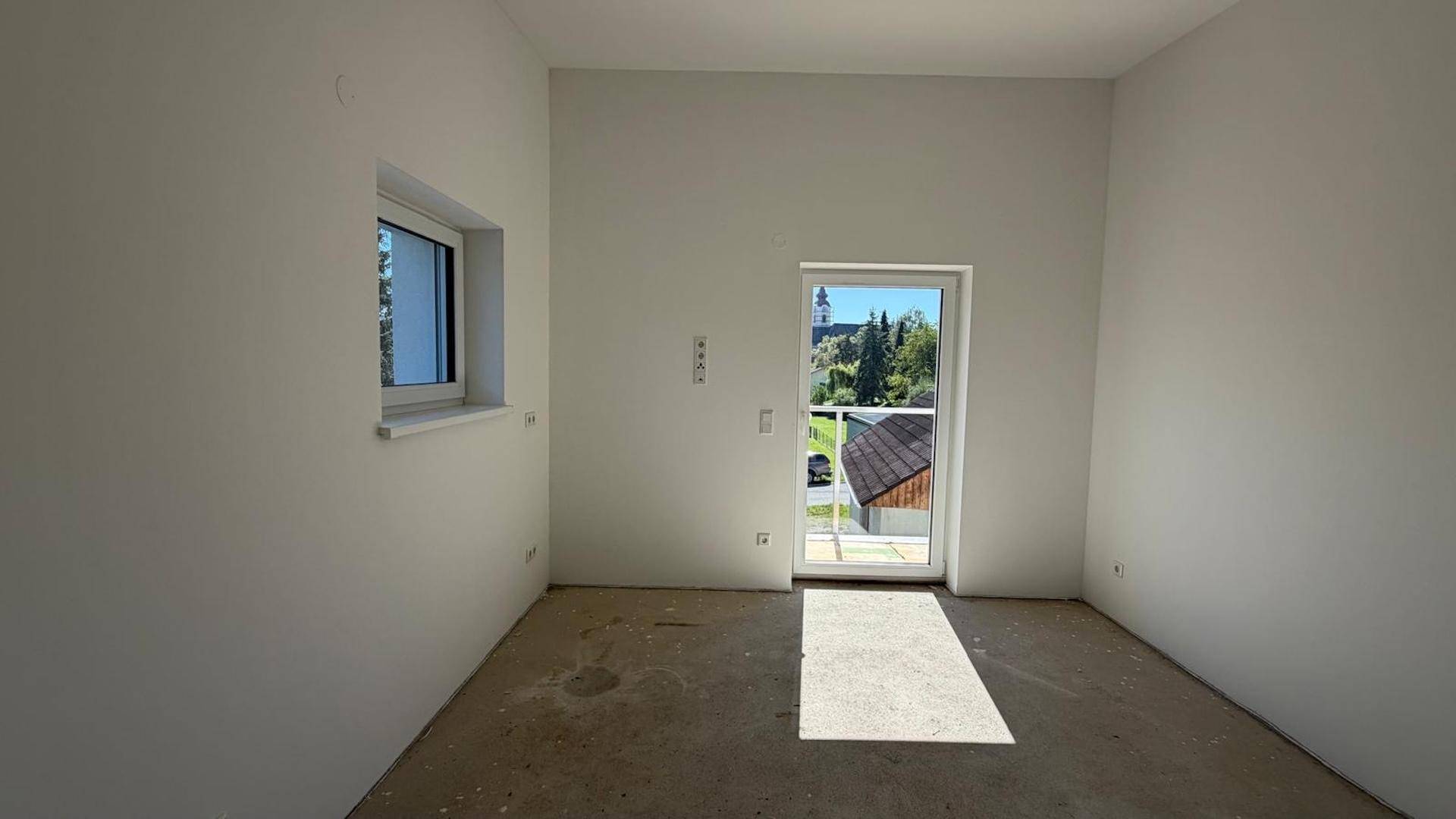 Heller Raum im Rohbau mit einer Tür zum Außenbereich und einem kleinen Fenster, das Licht spendet.