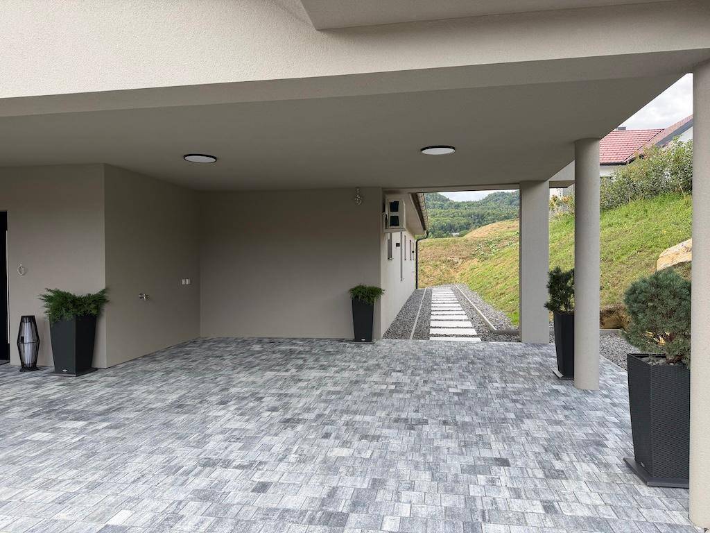 Blick durch den Carport auf einen gepflasterten Weg und die umliegende grüne Hügellandschaft des Grundstücks.