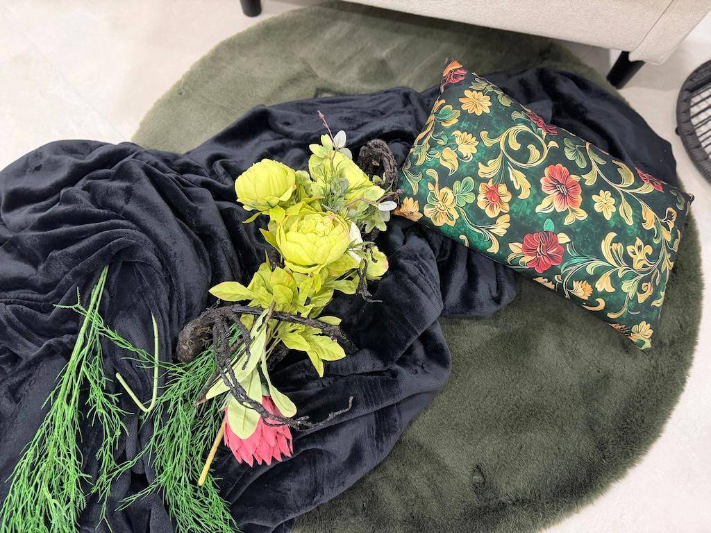 Dekorative Anordnung mit floralem Kissen, dunkler Decke und Kunstblumen auf einem grünen Teppich.