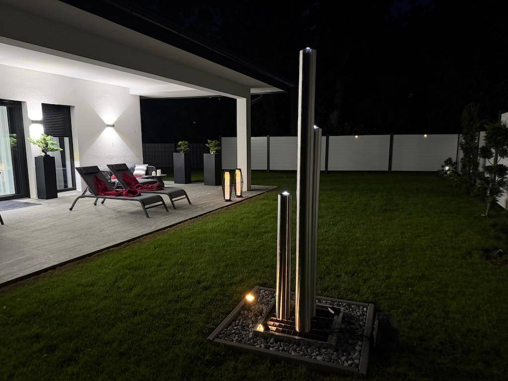 Abendliche Szene im Garten mit beleuchteter Terrasse, Liegestühlen und einem modernen Wasserspiel.