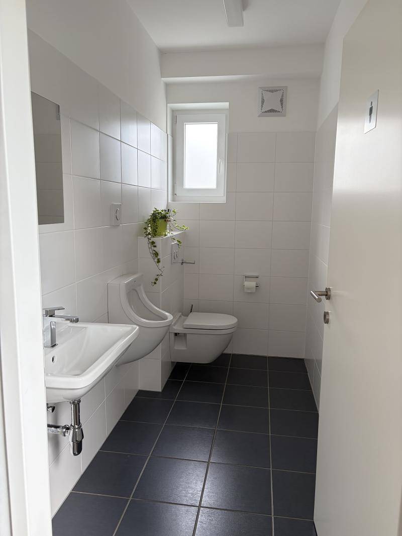 Helles Badezimmer mit Fenster, ausgestattet mit Waschbecken, WC und Urinal, ideal für gewerbliche Nutzung.