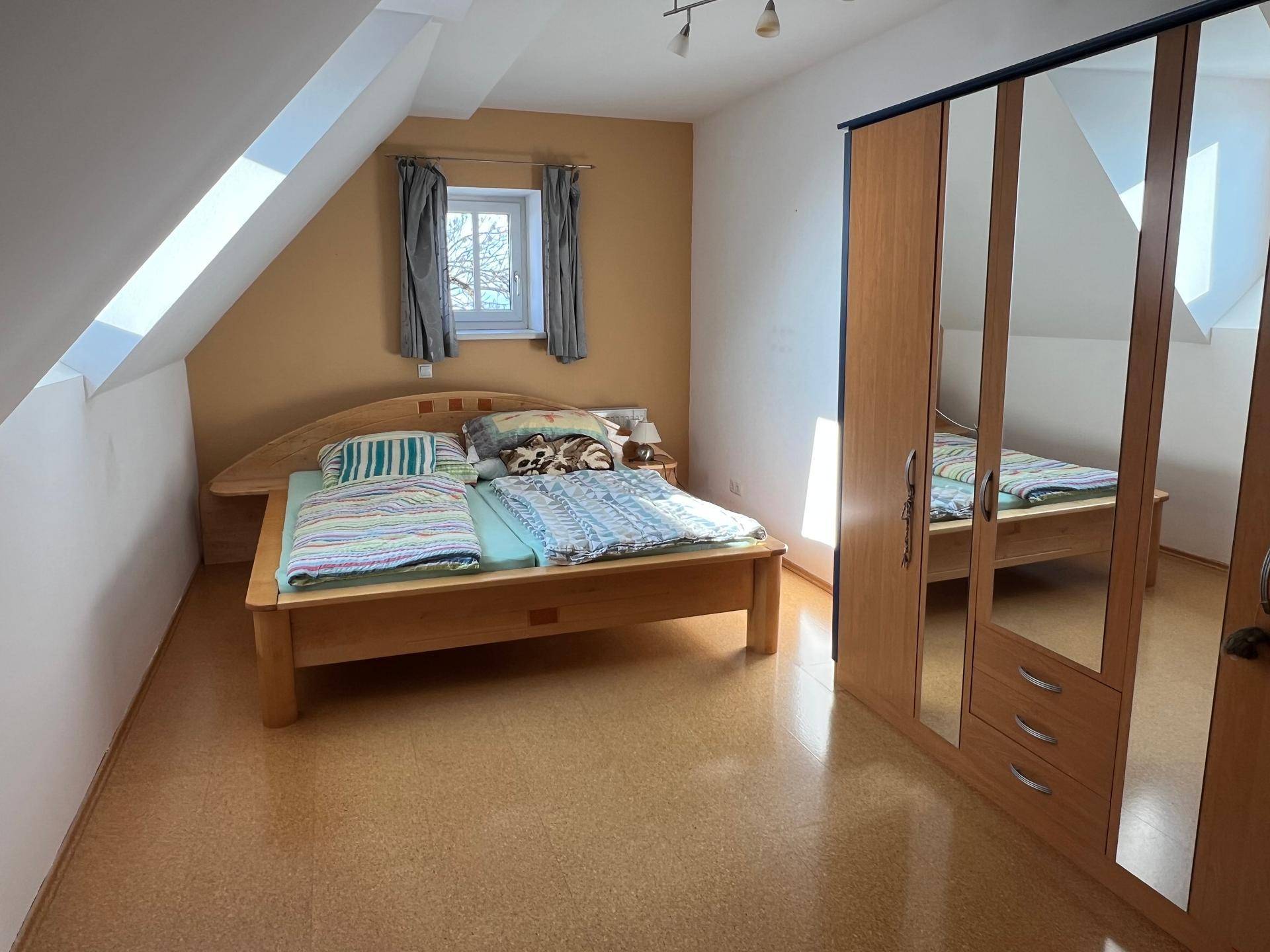 Schlafzimmer mit Holzbett, Spiegelschrank und Dachschräge mit kleinem Fenster.