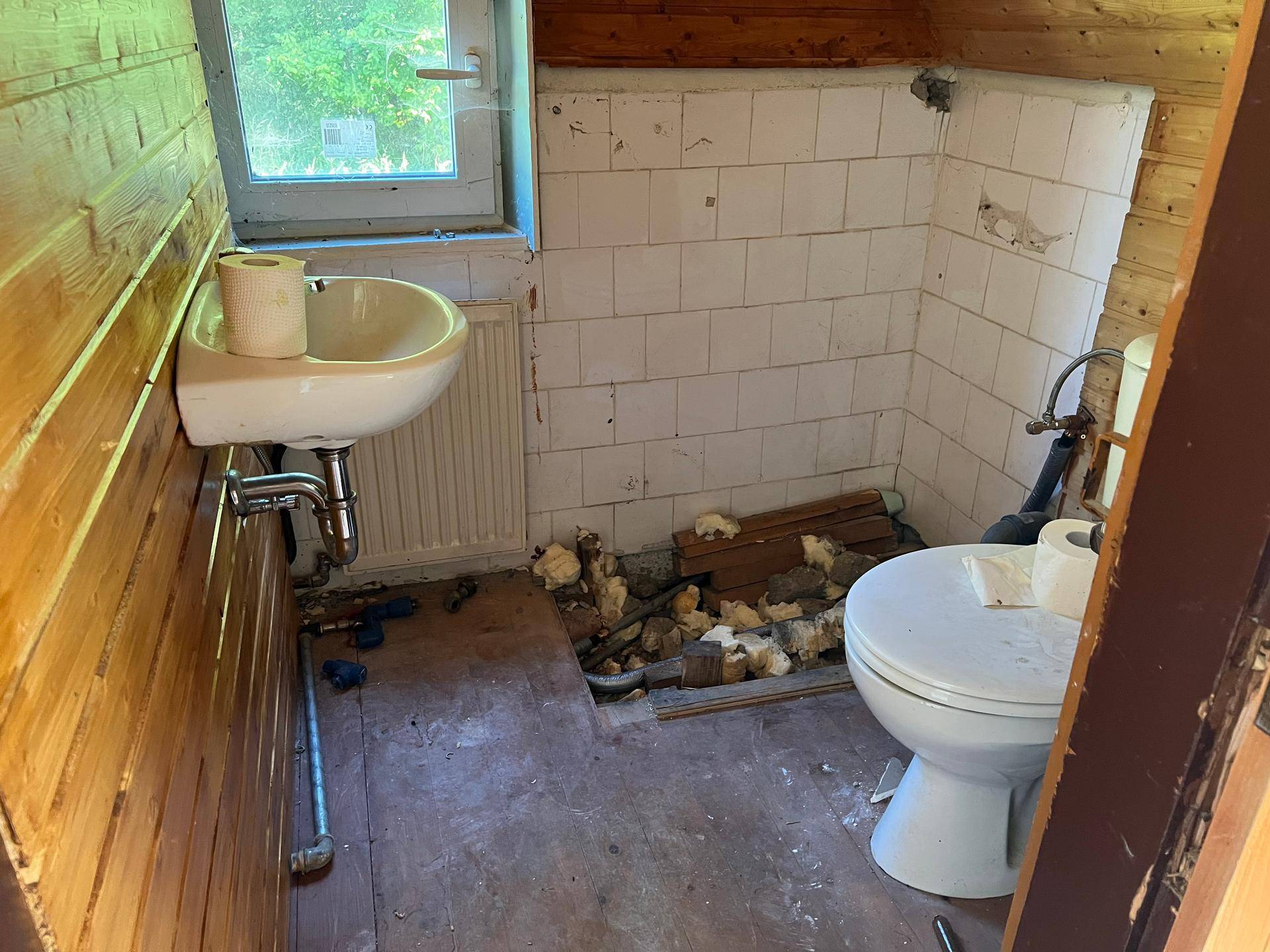 Einfache Toilette mit Holzverkleidung und Fenster, die Renovierungsbedarf zeigt.
