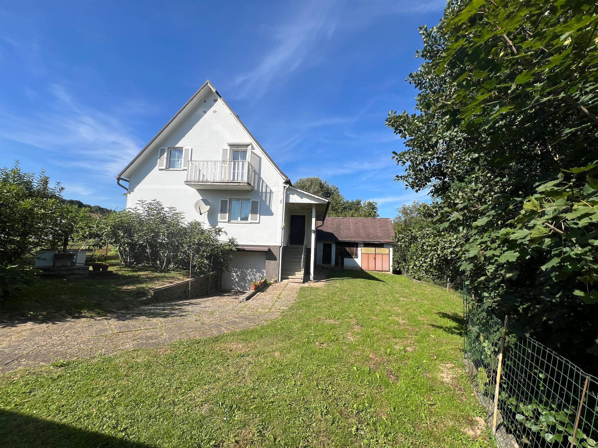 Freistehendes Haus mit Garten und Bäumen, umgeben von viel Grün unter blauem Himmel.