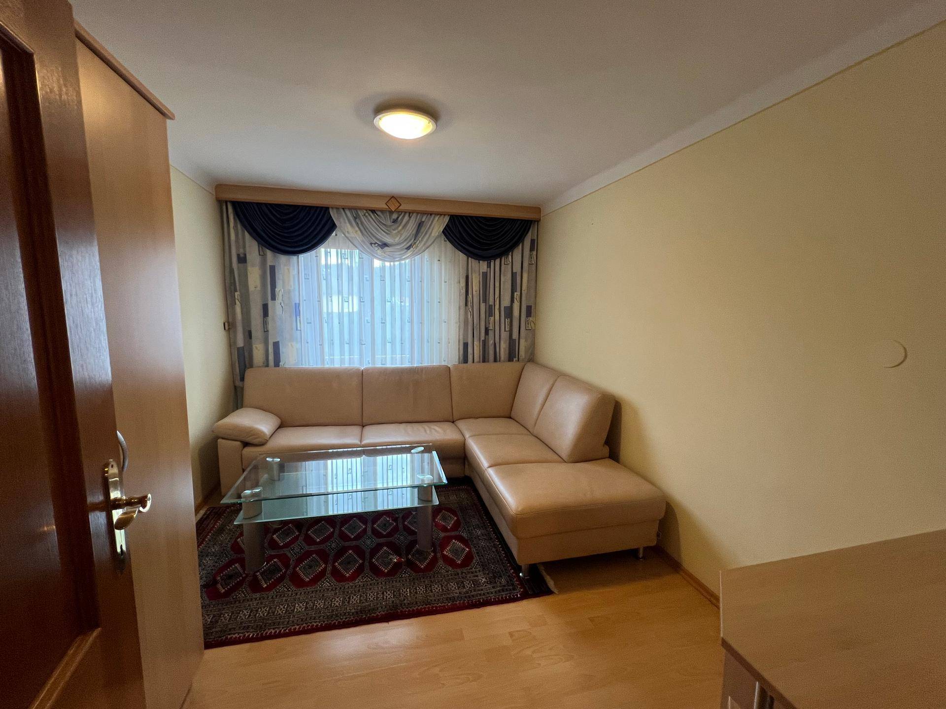 Wohnzimmer mit beiger Eckcouch, Glastisch und Parkettboden, dekoriert mit einem Teppich.