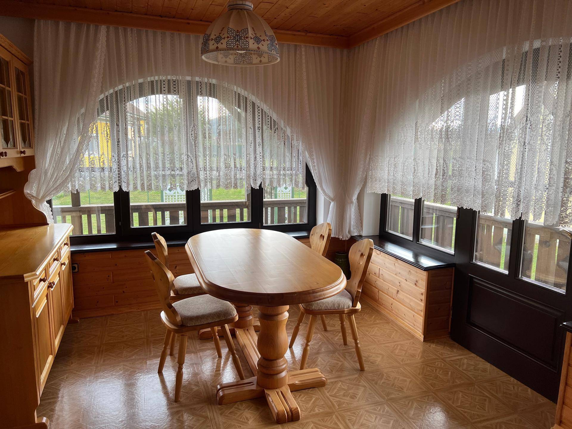 Heller Essbereich mit Holztisch und Stühlen, umgeben von großen Fenstern mit Blick ins Grüne.