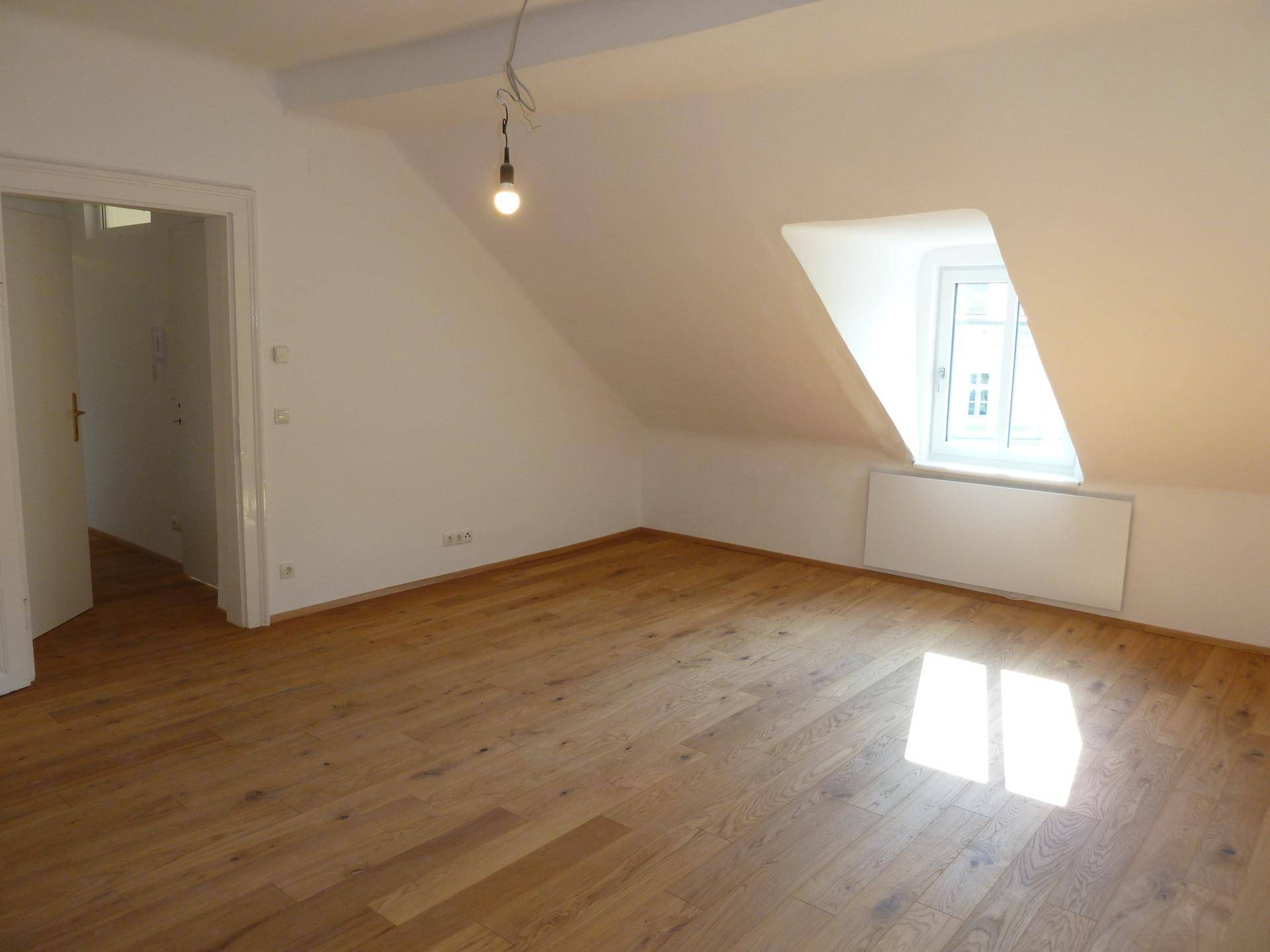 Heller Raum im Dachgeschoss mit schrägen Wänden, Holzboden und Fenster.