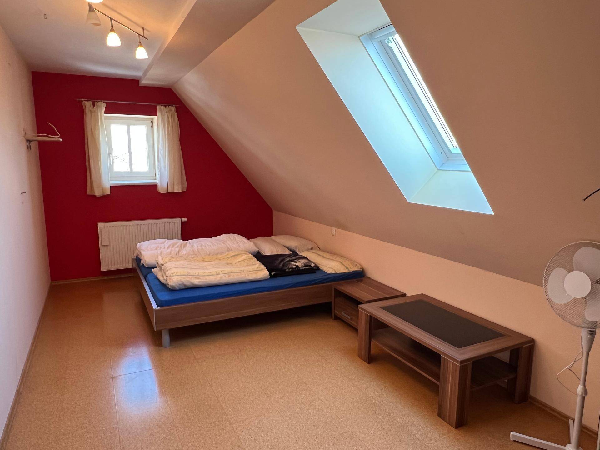 Helles Schlafzimmer mit Bett, Dachfenster und Dachschräge, betont durch eine rote Wand.