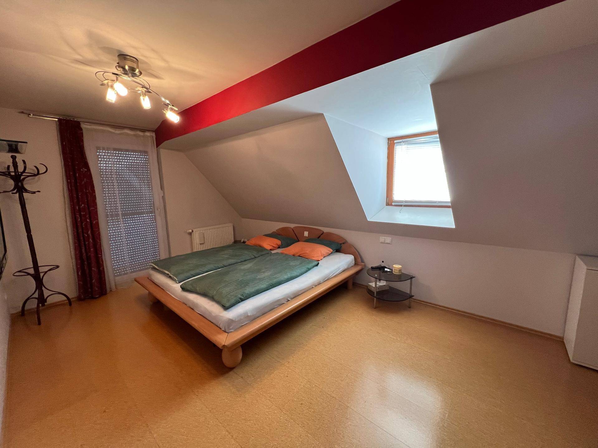Schlafzimmer mit Holzbett, kleinem Fenster und Dachschräge, akzentuiert durch eine rote Wand.