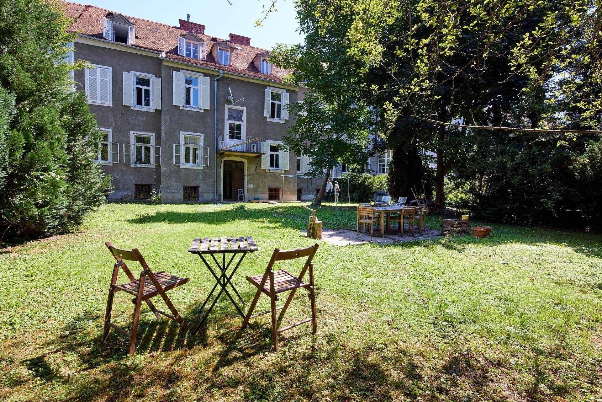 Gepflegter Garten mit Sitzgelegenheiten und altem Baumbestand vor dem Gebäude.