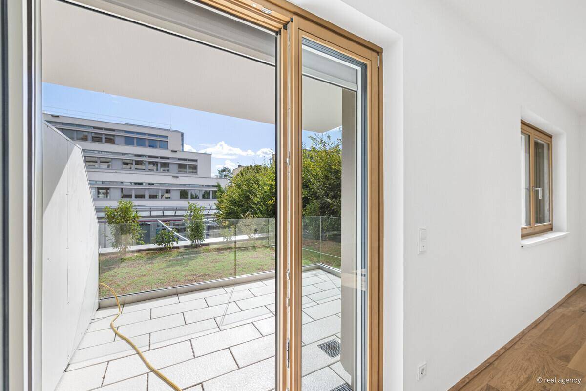 Blick von innen auf die geflieste Terrasse mit Glasgeländer und grüner Bepflanzung im Hintergrund.
