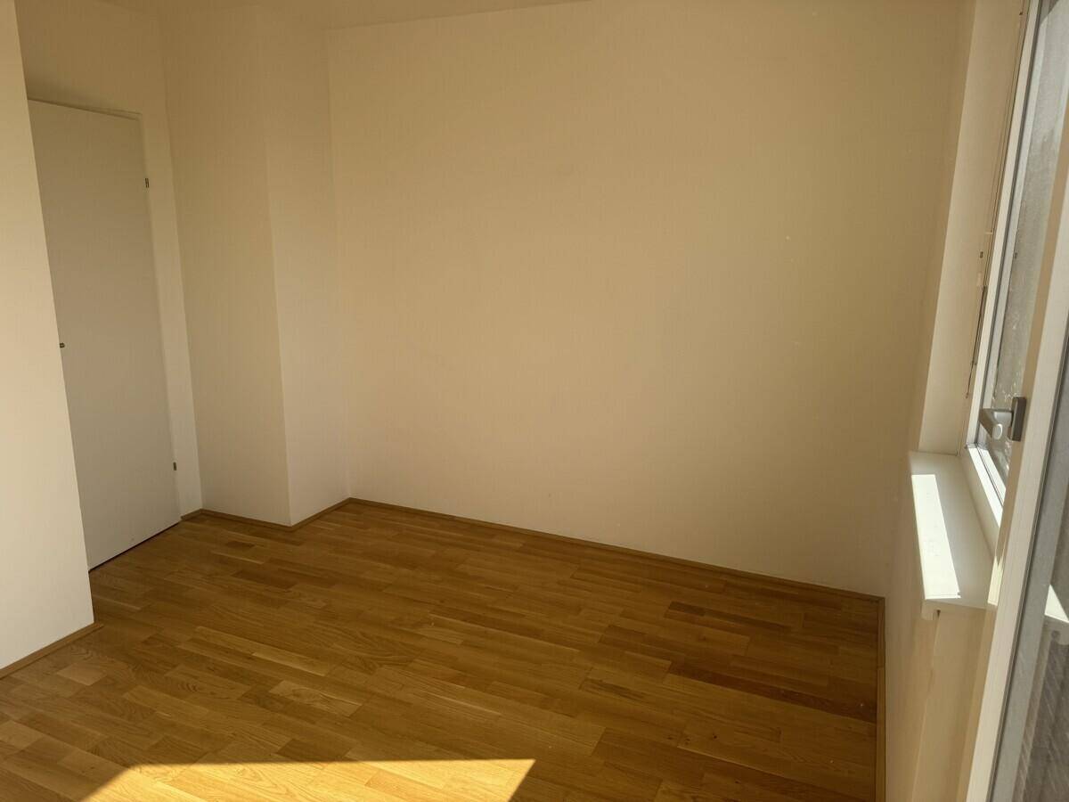 Leeres Zimmer mit Holzboden und einem Fenster, das Tageslicht hereinlässt.