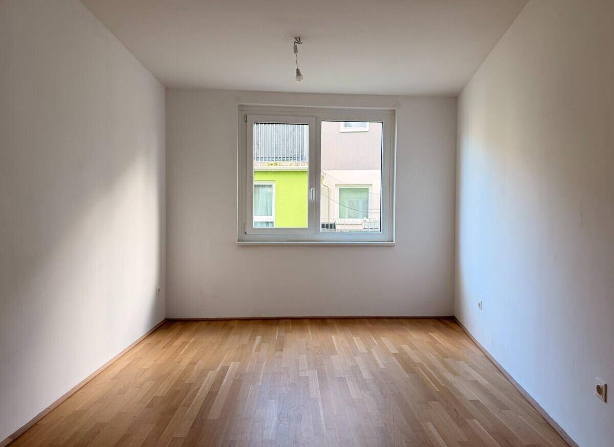 Leeres Zimmer mit Parkettboden und einem Fenster, das auf die gegenüberliegenden Gebäude blickt.