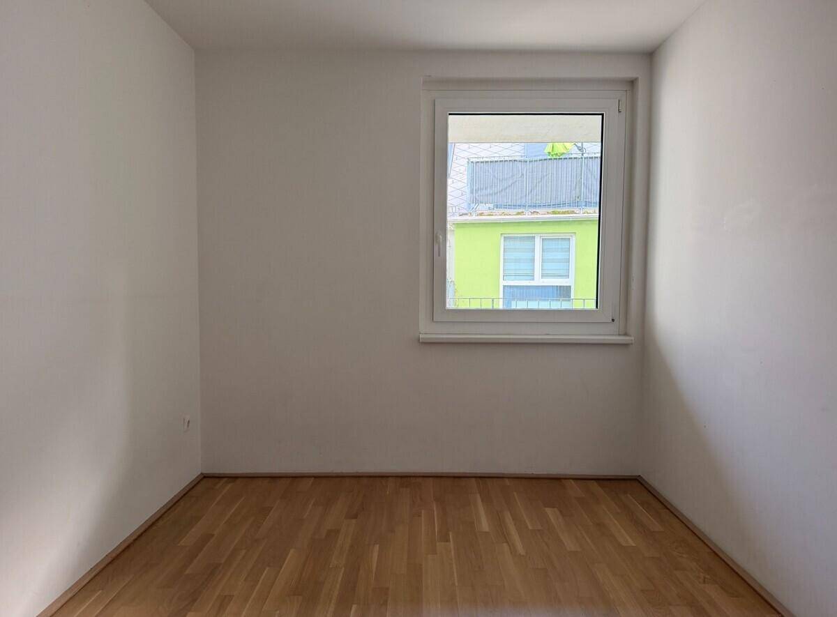Helles, leeres Zimmer mit Parkettboden und einem einzelnen Fenster, das Tageslicht hereinlässt.