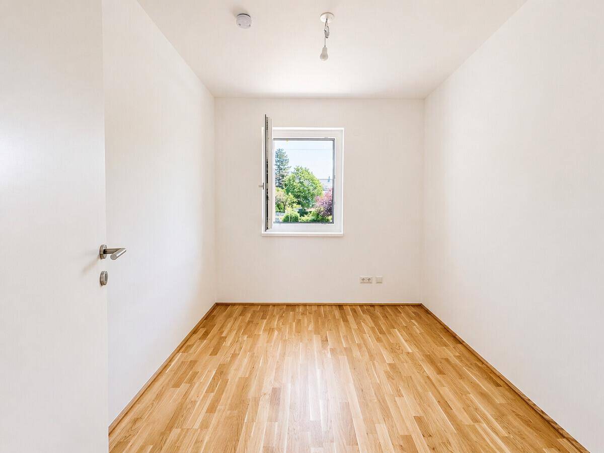 Ruhiges, helles Zimmer mit Parkettboden und Blick ins Grüne durch das Fenster.