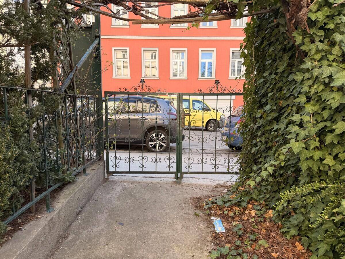 Blick durch ein verziertes Metalltor auf parkende Autos und ein gegenüberliegendes Gebäude mit roter Fassade in einer städtischen Umgebung.