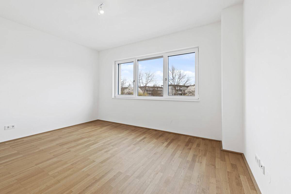 Helles, leeres Zimmer mit Parkettboden und breitem Fenster, bietet viel Tageslicht.