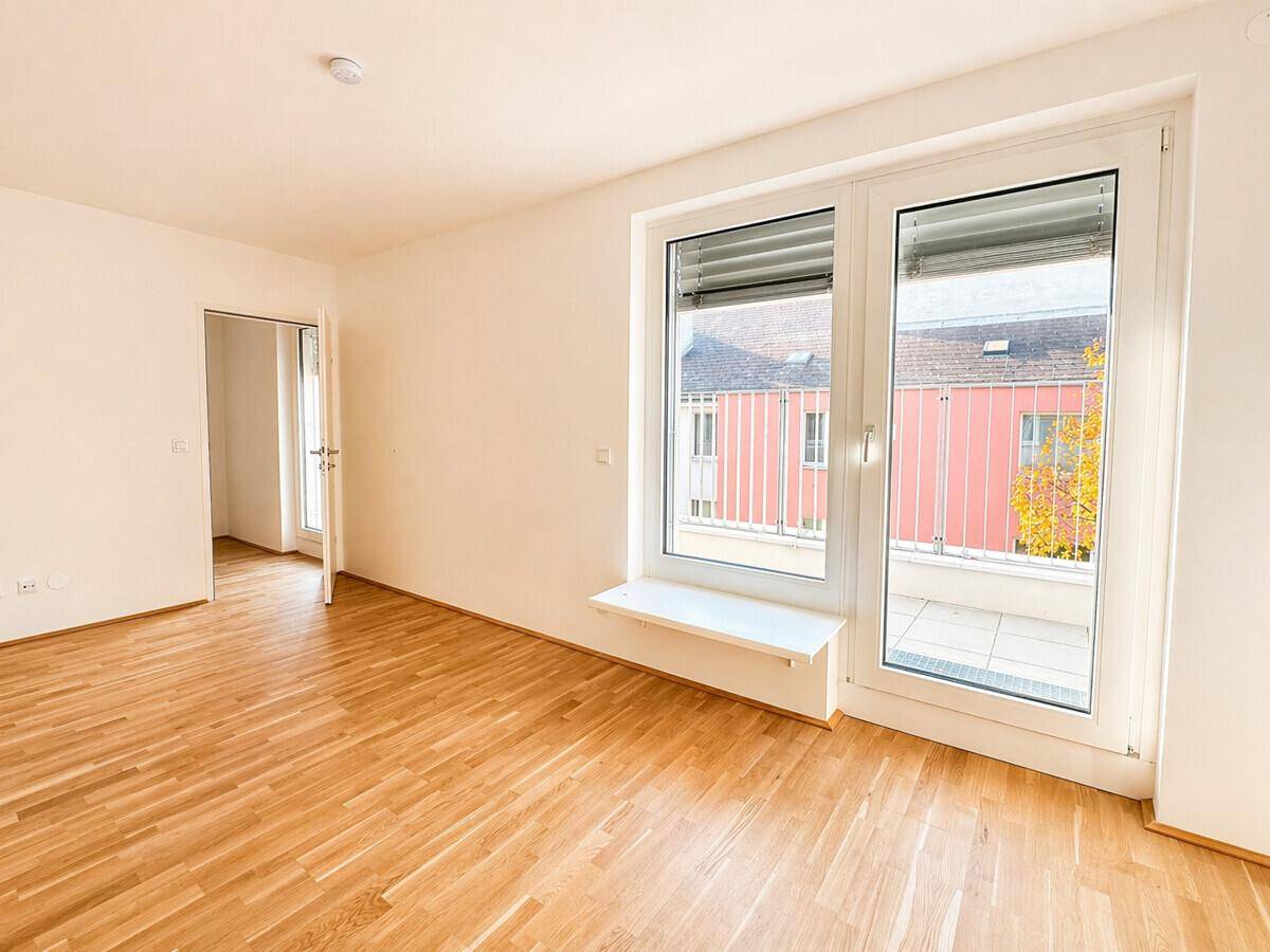 Geräumiges, helles Zimmer mit Holzboden und großen Fenstern, die auf einen Balkon führen.