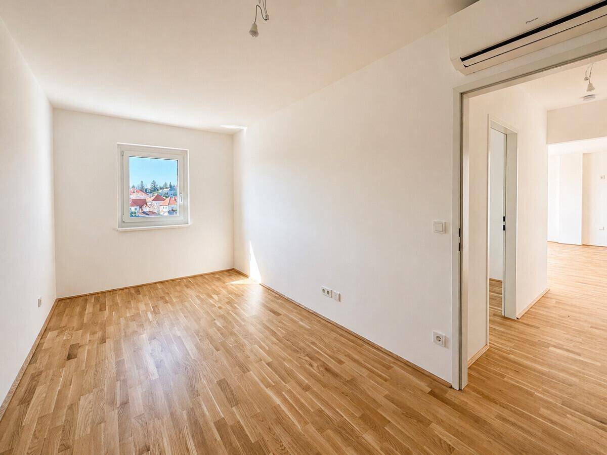 Leeres Zimmer mit Parkettboden, einem Fenster und einer Klimaanlage, bereit zur individuellen Gestaltung.
