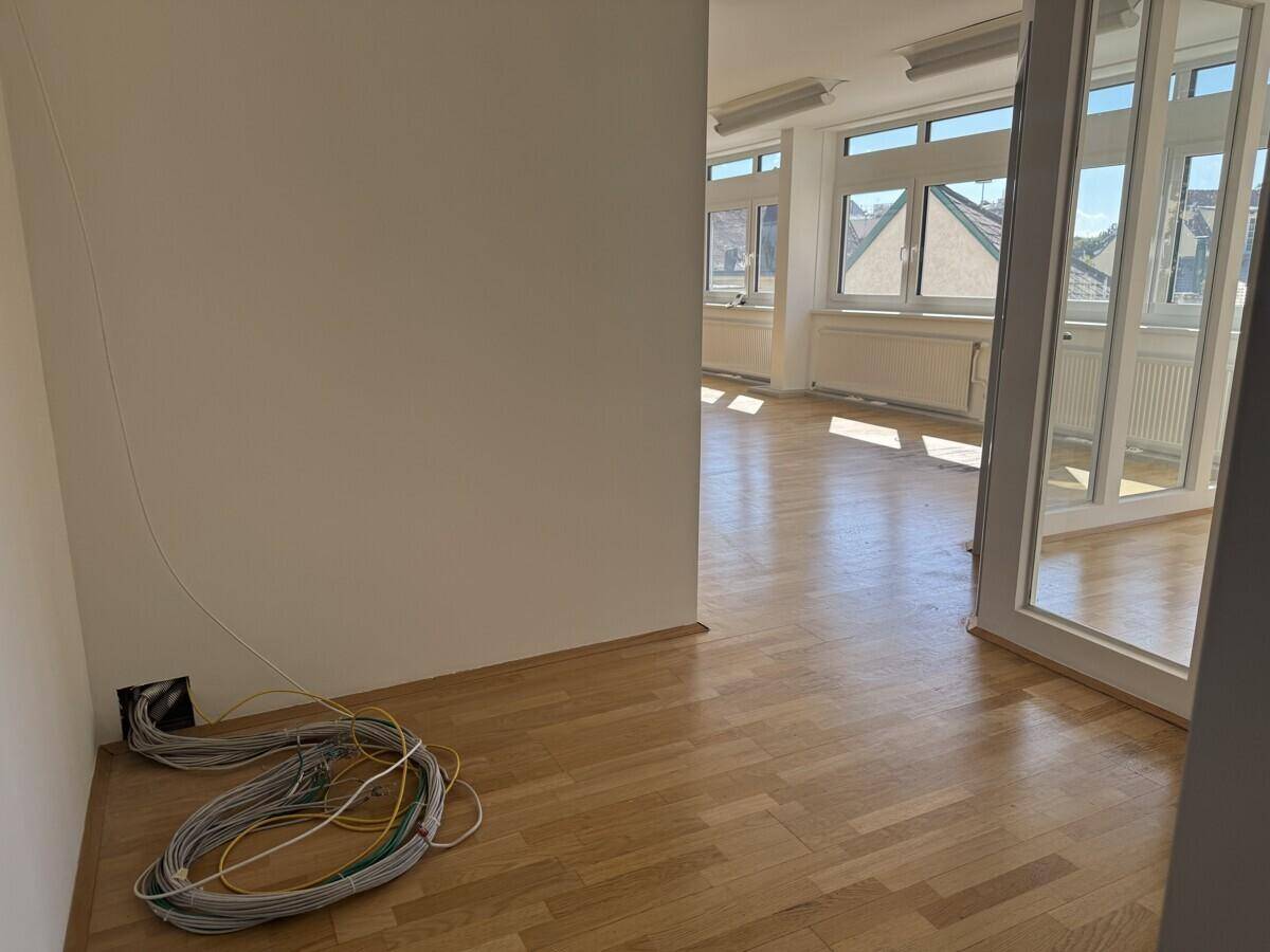 Ecke eines Büroraums mit sichtbaren Kabeln und einem Blick durch eine Glastür in einen weiteren hellen Bereich.