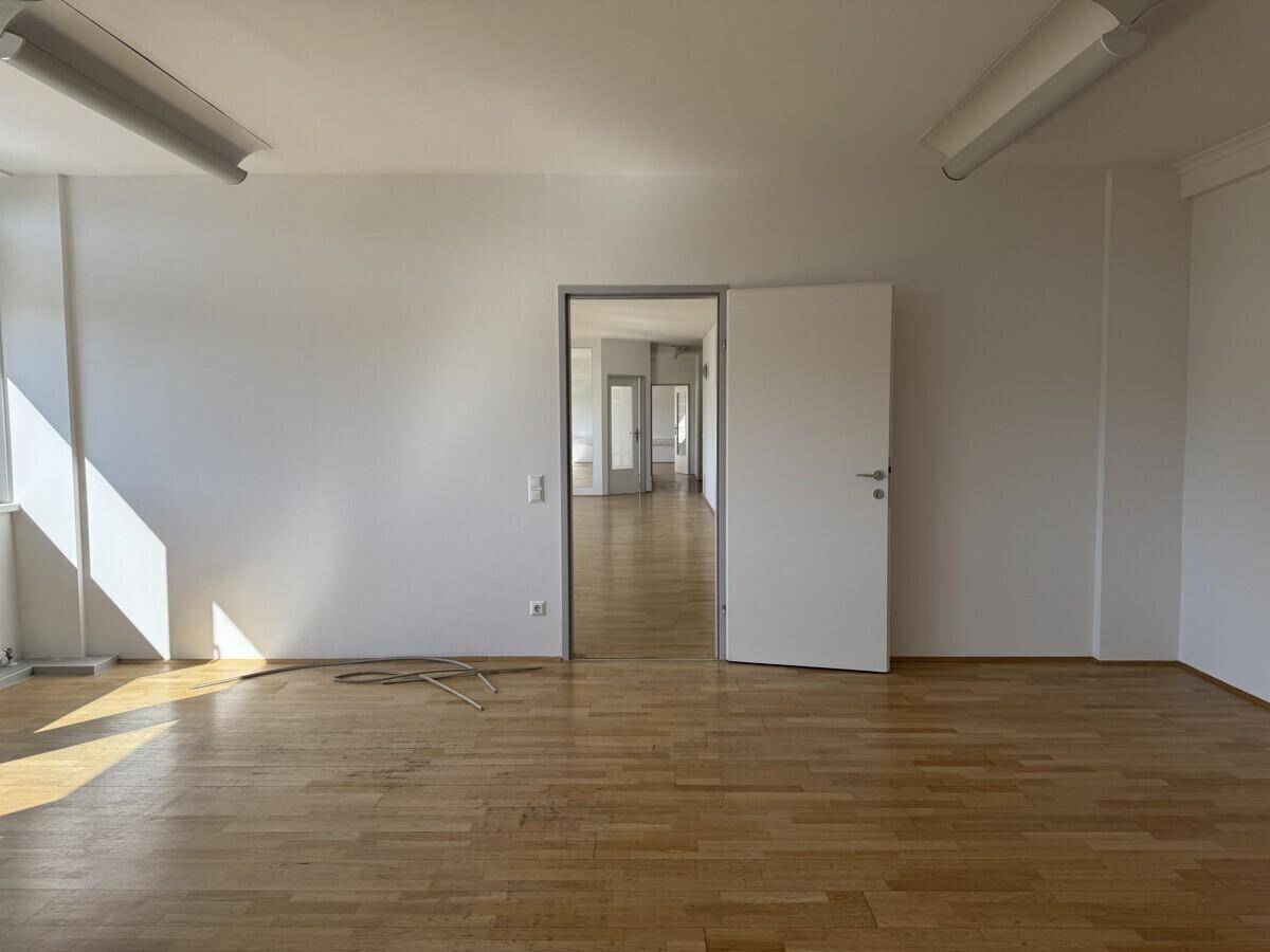 Leerer Büroraum mit Holzboden und weiß gestrichenen Wänden, der zu einem langen Flur führt.