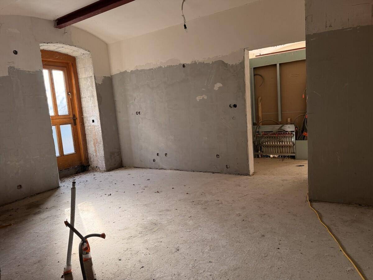 Großer Raum im Rohbau mit unverputzten Wänden, einem Holztürrahmen und Blick in einen angrenzenden Bereich mit weiteren Installationen.