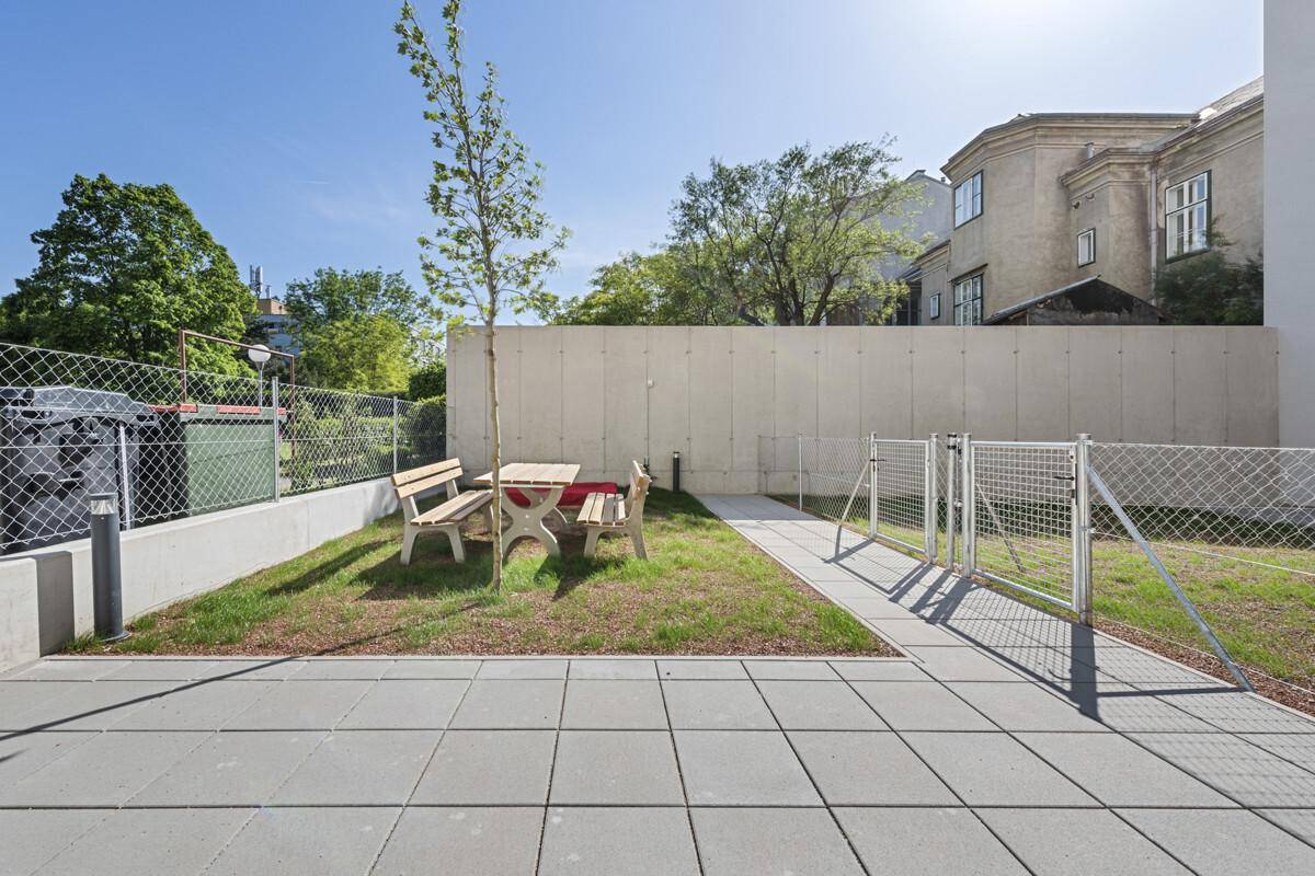 Gemeinschaftlicher Gartenbereich mit Sitzgelegenheiten und einem jungen Baum unter blauem Himmel.